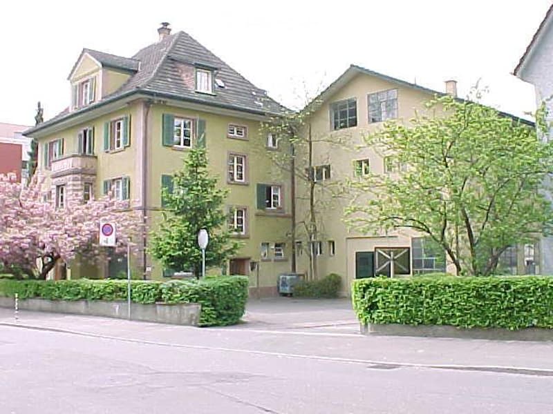 Two-story residential building with yellow facade, green shutters, and multiple windows. The building features a pitched roof and is surrounded by greenery and a low hedge along the sidewalk.