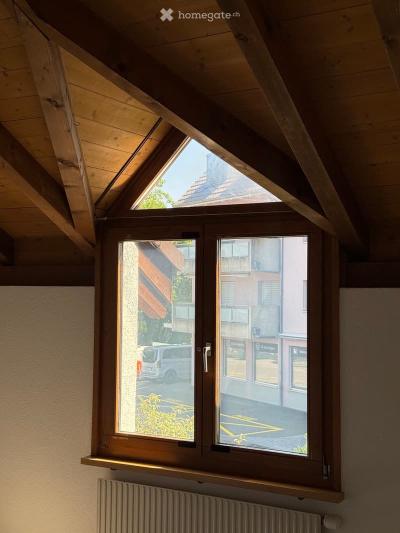 An attic room with wooden slanted ceilings, large windows showing a view of the street and neighboring buildings, a radiator, and a wooden shelf below the windows.