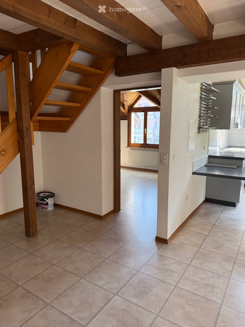 Empty hallway with wooden staircase, tiled floor, and a view into a kitchen area with modern cabinets and a countertop.