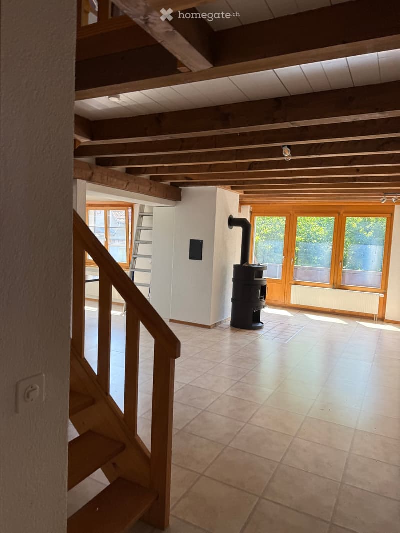 Empty room with exposed wooden beams, tiled floor, staircase, and a wood-burning stove. Natural light from large windows and a door.
