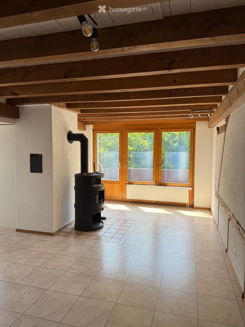 Bright hallway with exposed wooden beams, tiled floor, and a black wood-burning stove on the left side.