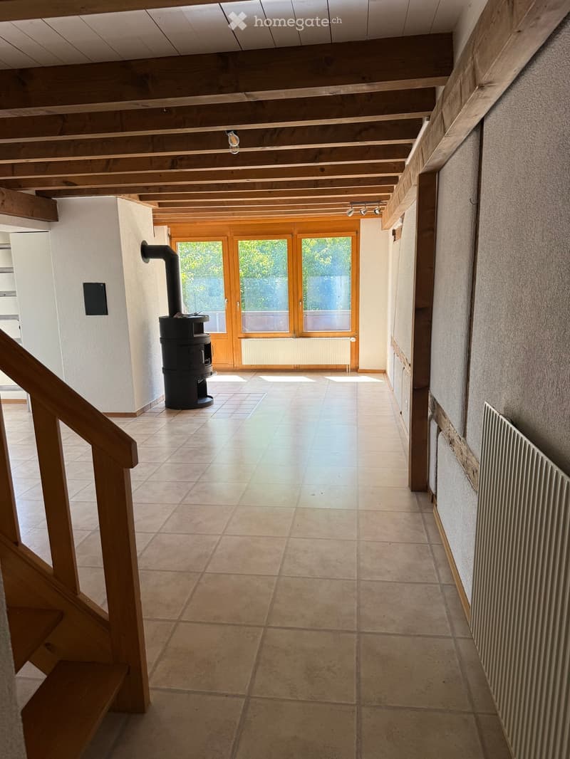 Hallway with tiled floor, wooden ceiling beams, staircase on the left, wood-burning stove, large glass doors leading outside.