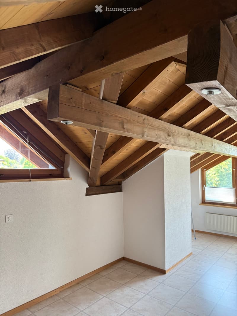 An unfurnished room with exposed wooden beams and a slanted wooden ceiling, featuring tiled flooring and large windows with wooden frames.