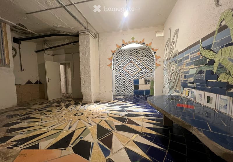 Corridor with colorful mosaic tiles on walls and floor, arched doorway, exposed pipes and lighting fixtures on the ceiling.