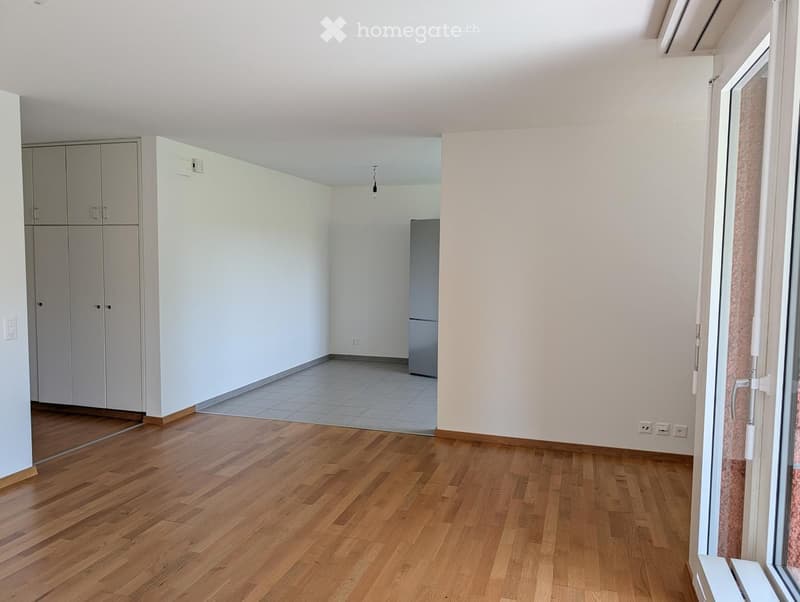Empty room with wooden flooring, white walls, and a large window with white frame. There is a small tiled area in the corner, and a set of white cabinets is visible on the left side.