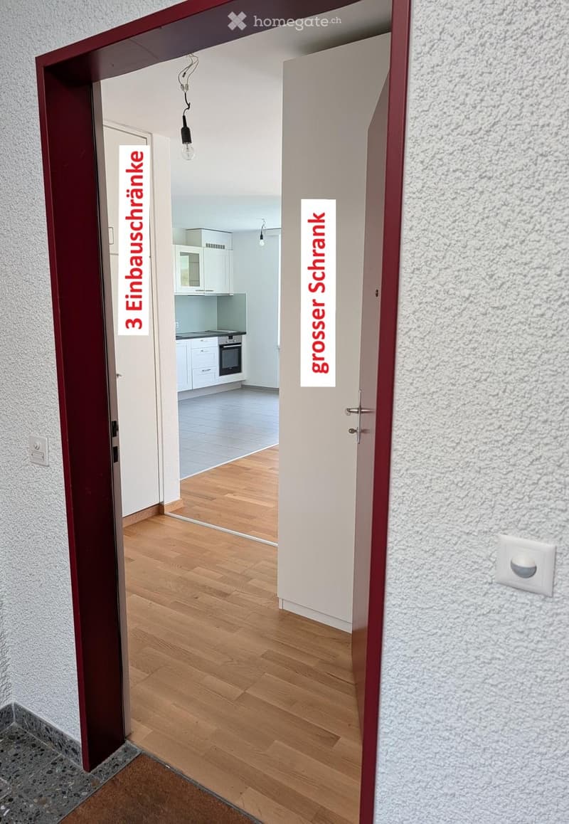 Hallway with wooden flooring, leading to a kitchen area with white cabinets and appliances.