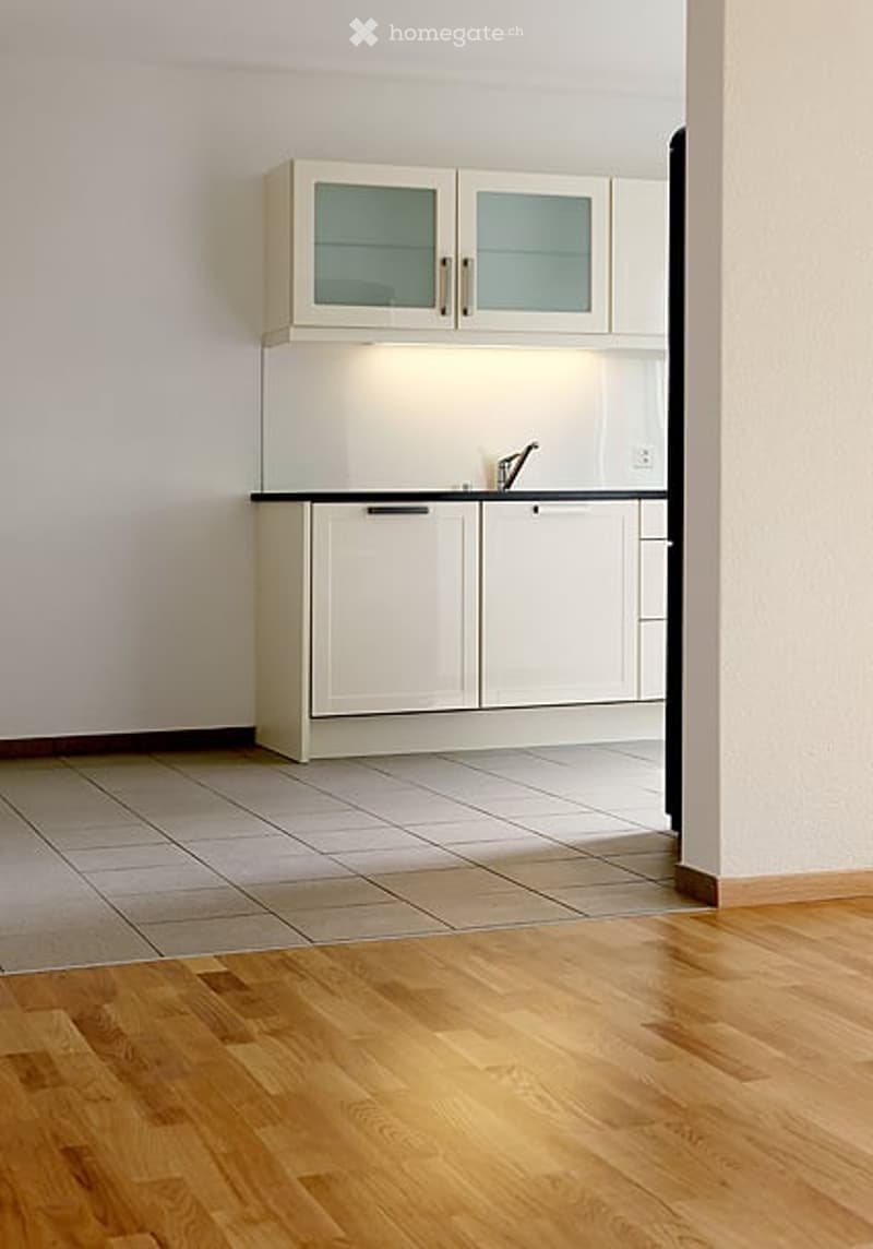 Modern kitchen with white cabinets, glass-fronted upper units, and a sleek black countertop. Wooden flooring and bright lighting.