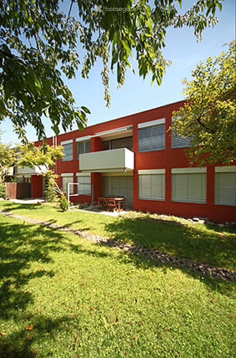 Red brick building with white trim, multiple windows, and a small balcony. The surrounding area includes a grassy lawn and a small patio with a table and chairs.