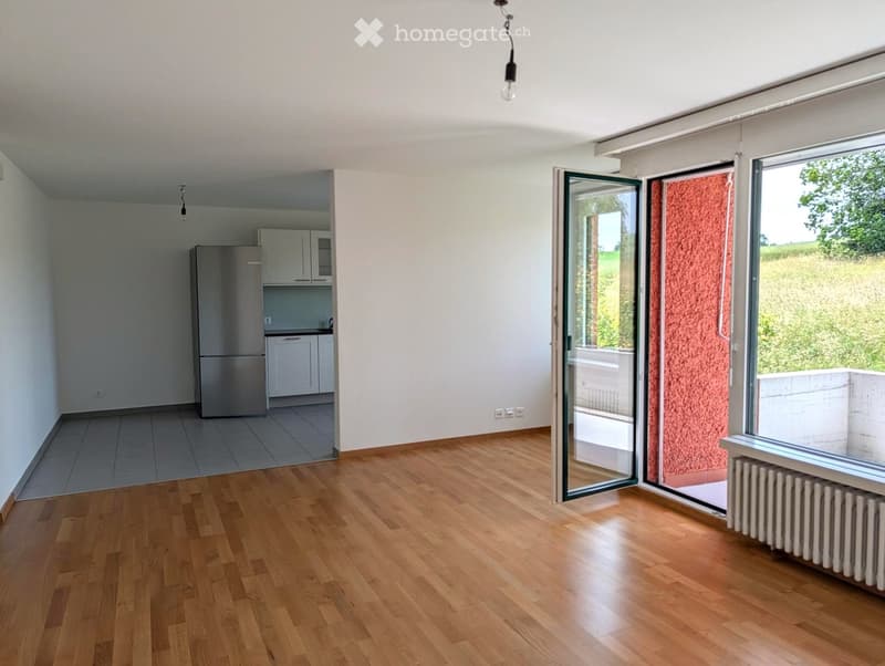 Bright, open living space with wooden flooring, large sliding glass doors leading to a balcony, and a small kitchen area with white cabinets and a stainless steel refrigerator.