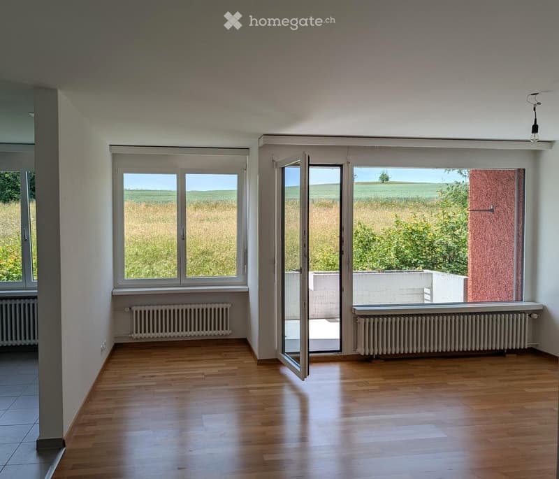 Empty room with wooden flooring, large windows, radiators, and an open sliding door leading to a balcony or outdoor area.