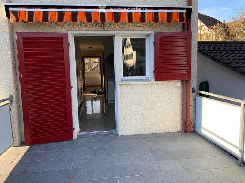 Balcony with tiled floor, metal railing, and an open doorway leading into a room with visible furniture.