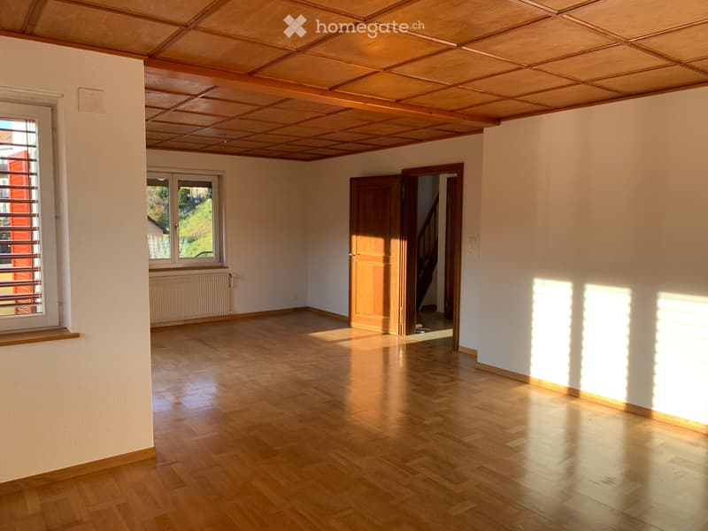 Empty room with wooden flooring, white walls, and large windows allowing natural light. Features a wooden ceiling and a door leading to another area.