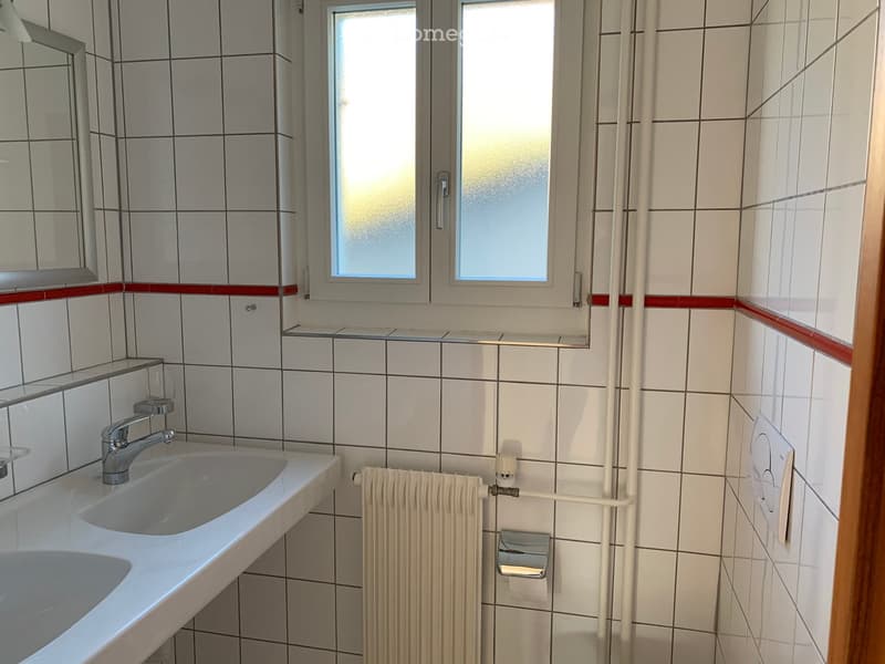Clean bathroom with white tiled walls, a single sink with a modern faucet, a radiator, a mirror, and a toilet paper holder. The window has frosted glass panes.