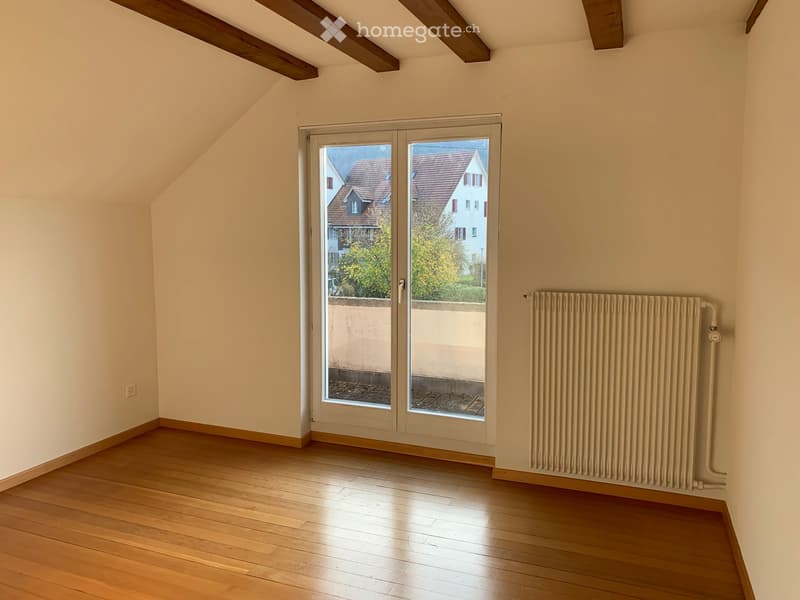 Empty attic room with wooden flooring, exposed wooden beams, large sliding glass door leading to a balcony, and a radiator on the wall.
