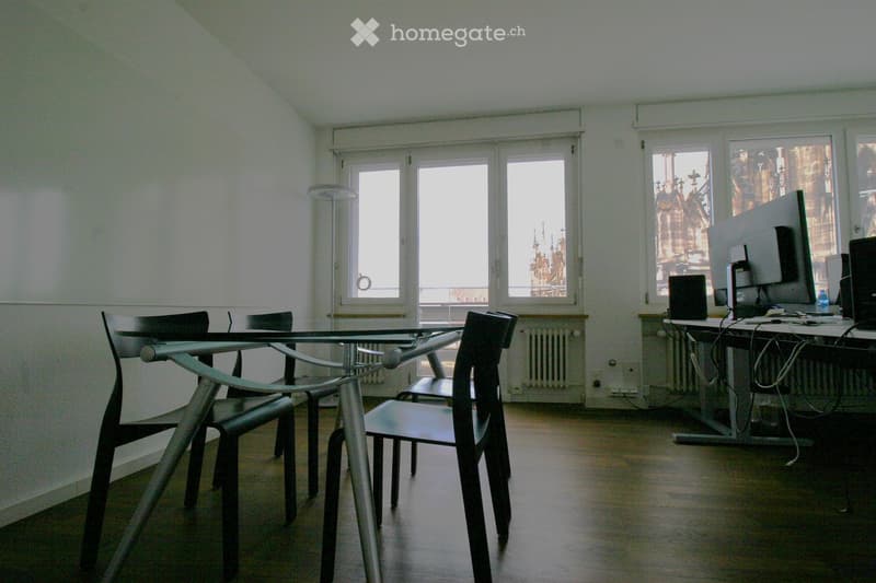 A modern, minimalist room with a glass table and chairs, a computer setup with a large screen, and large windows offering a view of a historic building.