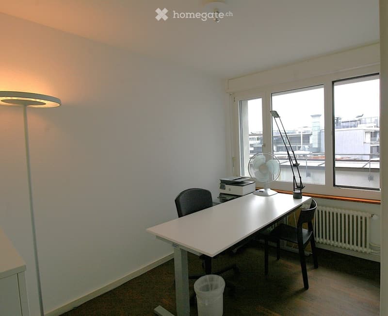 A small, well-lit room with a white table, two chairs, a fan, a printer, a lamp, and a radiator near the window.