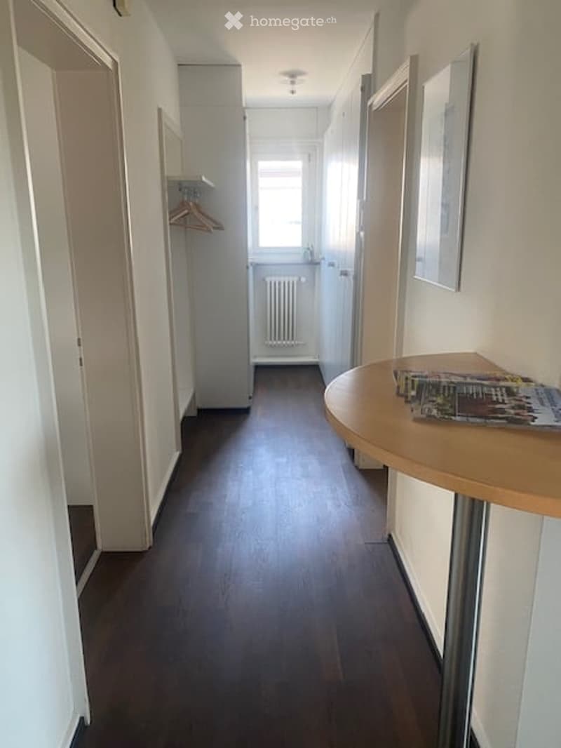 A narrow hallway with wooden flooring, white walls, and a round table with magazines on it. There is a coat rack with hangers and a radiator visible in the background.