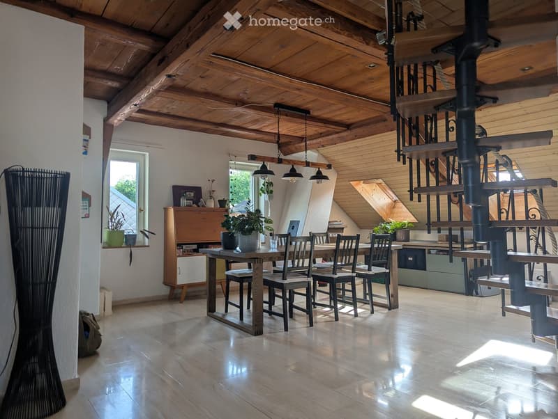 Open-plan living and dining area with wooden beams, exposed wooden ceiling, large windows, dining table with chairs, cabinet, potted plants, and a laptop on the table.