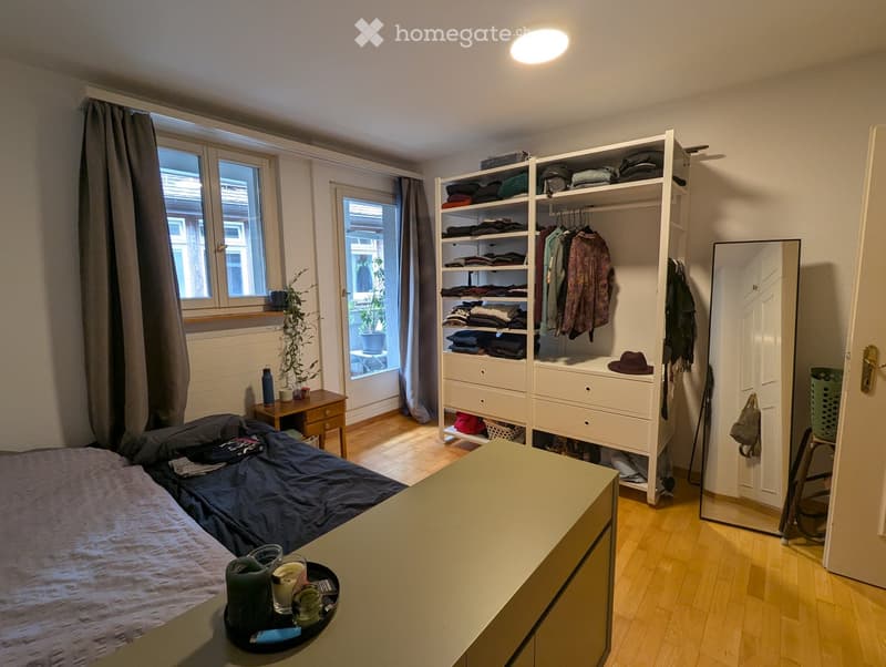 Cozy bedroom with a bed, nightstand, wardrobe, and a small table with a plant and candles. The room features wooden flooring and large windows with curtains.
