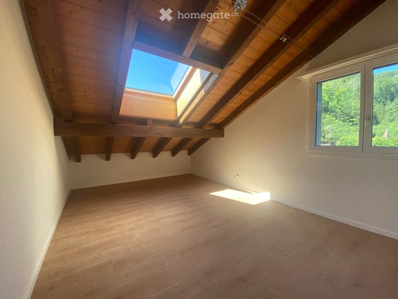 Empty attic room with wooden floor, exposed wooden beams, skylight windows, and a large window showing a green view.