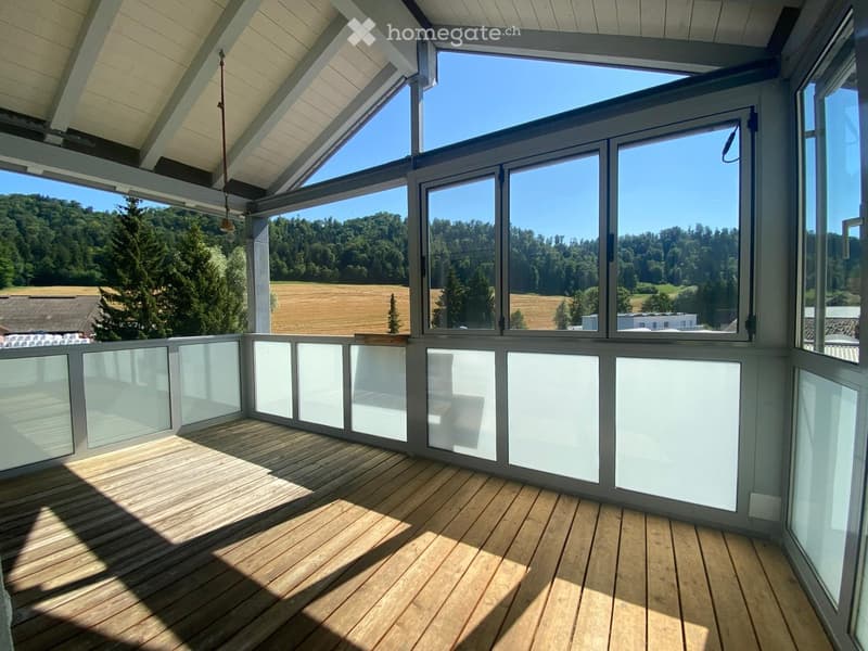 Bright, spacious balcony with wooden flooring, large glass windows, and a scenic view of a green landscape and fields.