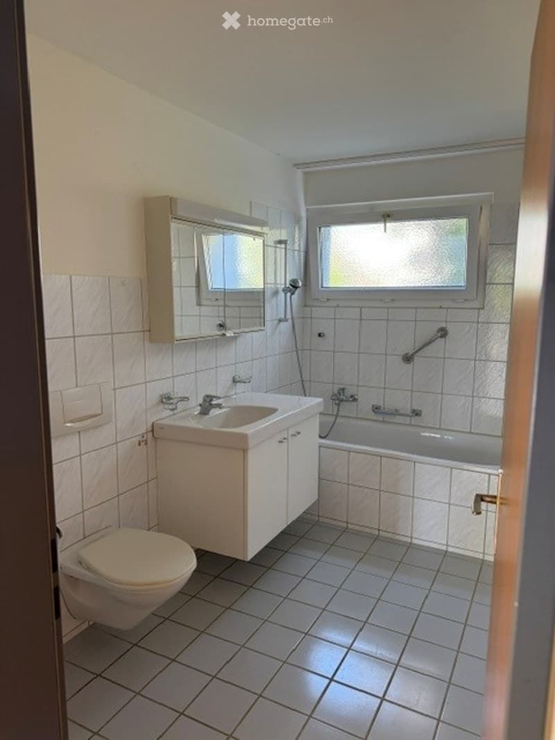 Modern bathroom with white tiles, a sink, toilet, bathtub, and a mirror above the sink. The room features a window with frosted glass and a grab bar next to the bathtub.