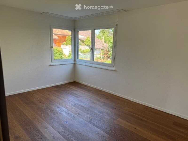 Completely empty room with wooden flooring and large windows, showing a view of the garden and neighboring houses.
