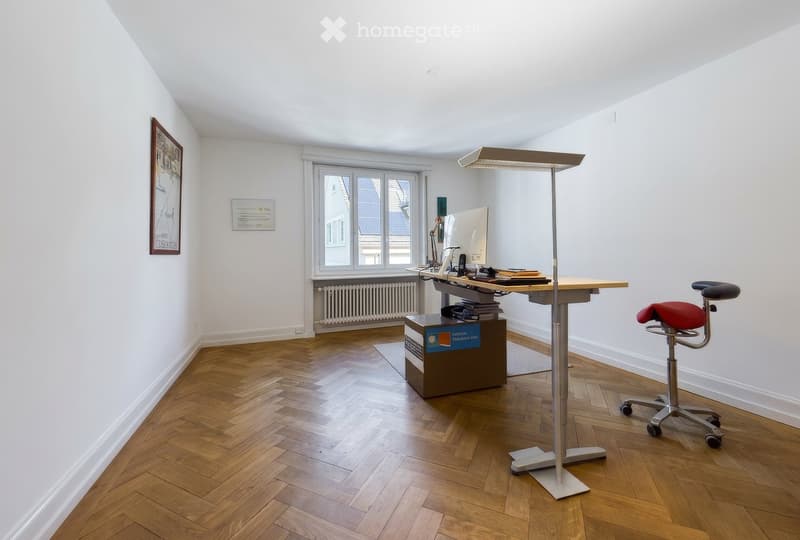 Bright, modern office space with wooden herringbone flooring, white walls, large window, adjustable standing desk, ergonomic chair, computer monitor, and a floor lamp.