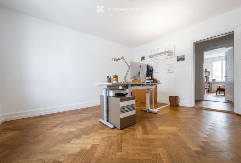 Modern office space with wooden flooring, white walls, and a standing desk setup. Includes a computer, printer, chair, lamp, small plant, and picture frames on the wall. An open doorway leads to another room with visible furniture.