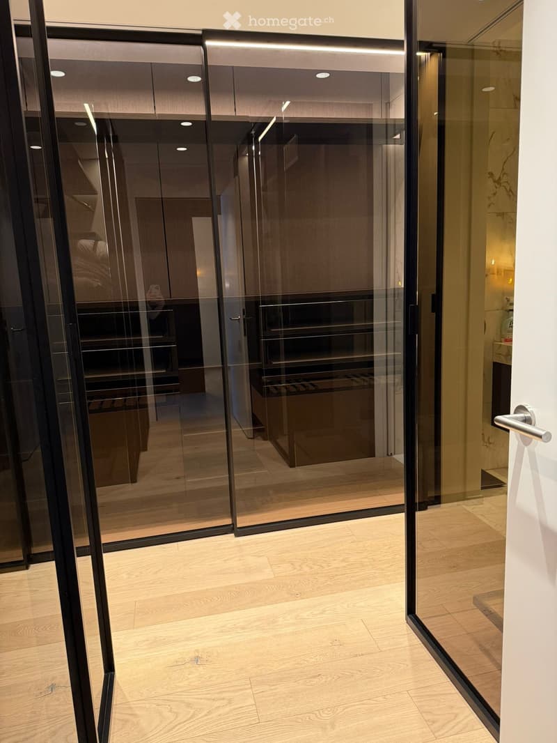 Modern hallway with light wooden flooring, glass sliding doors leading to a closet area, and a glimpse of a bathroom with marble countertops.