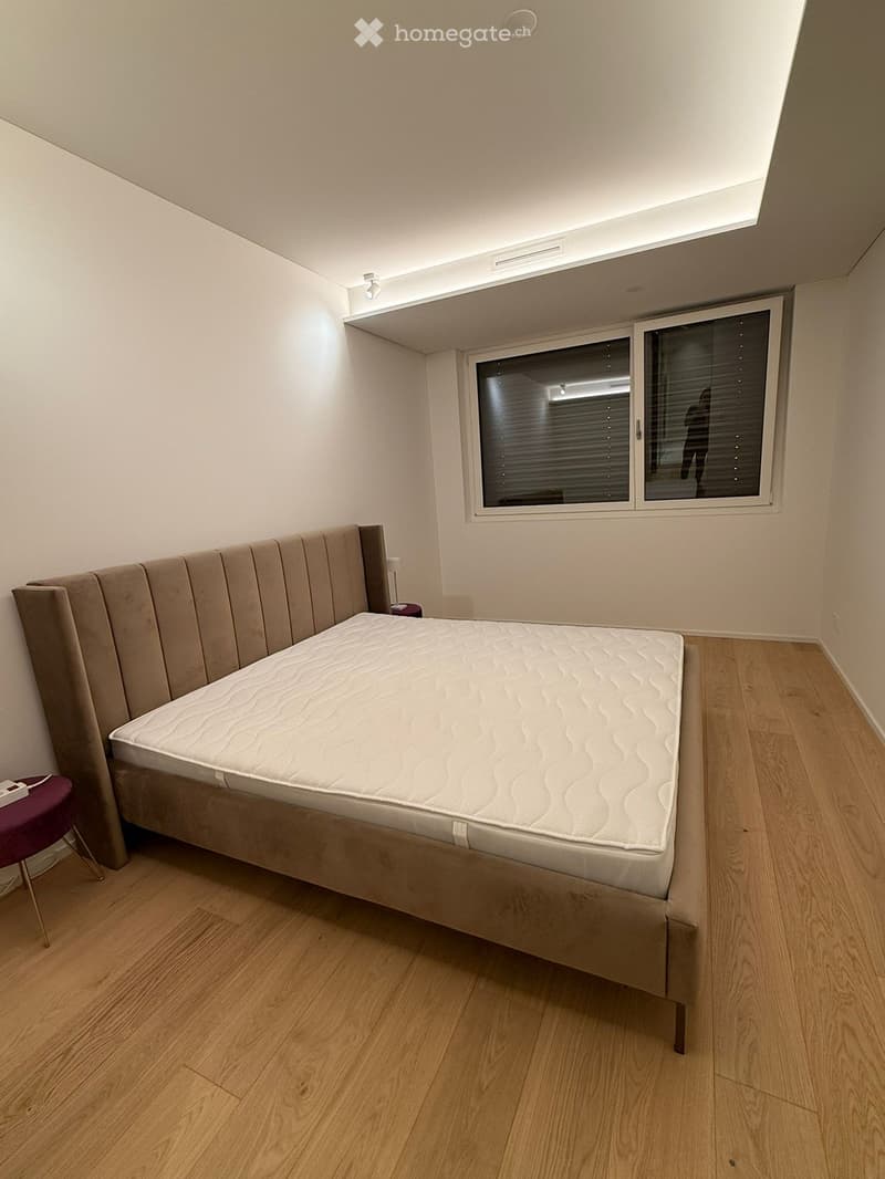 Modern bedroom with a large bed, upholstered headboard, and a small purple stool near the window. The room features wooden flooring and large windows with blinds.