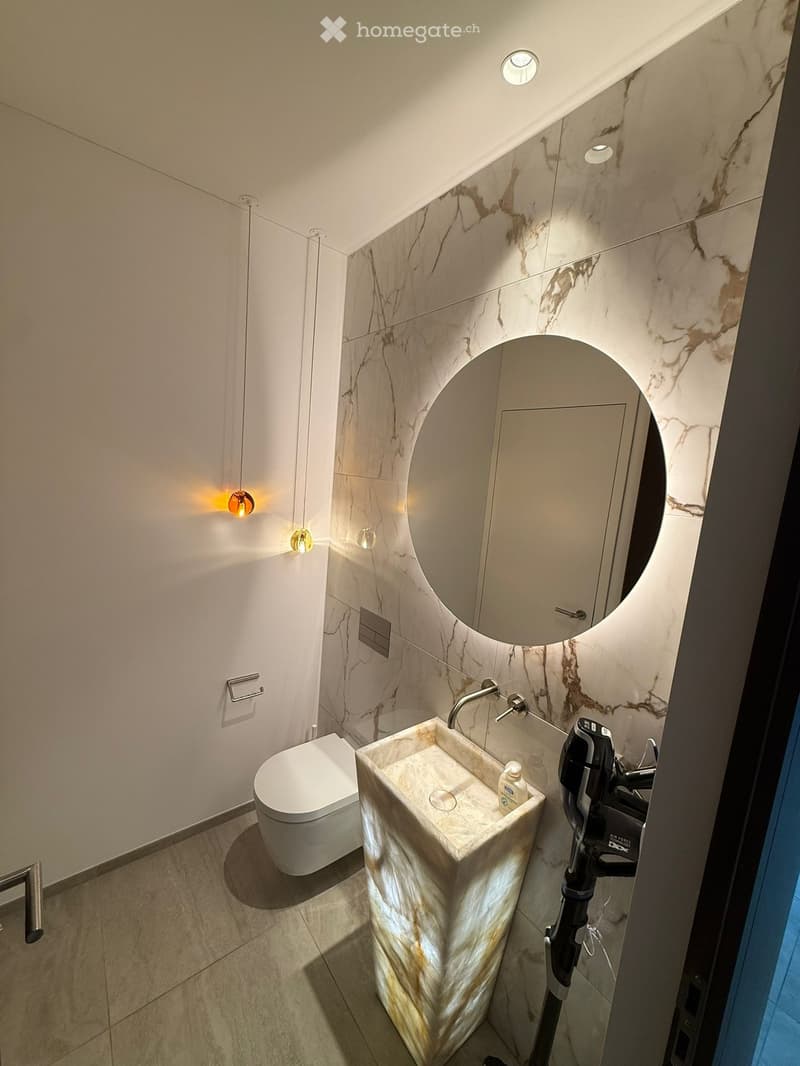 Modern bathroom with marble walls, a white toilet, a marble sink, a large round mirror, and wall-mounted lighting.