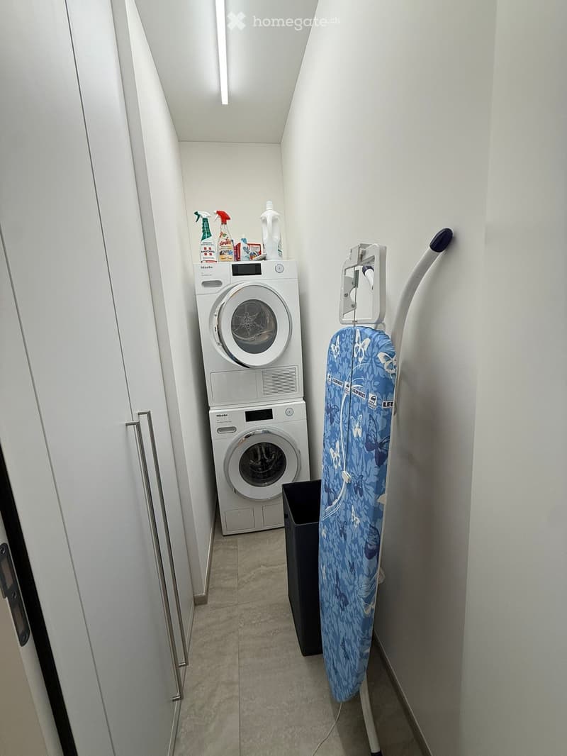 A small, well-lit room with a washing machine, an ironing board, and cleaning supplies on a shelf.