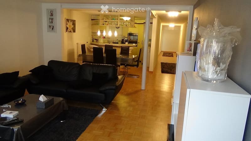A modern living room with a black leather sofa, coffee table, and a dining area with chairs and a table. The room features wooden flooring and a hallway leading to a kitchen area.