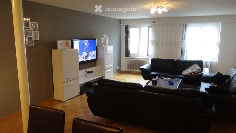 Modern living room with wooden flooring, large windows, black leather sofa, coffee table, TV cabinet, and a couple of chairs.