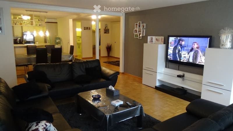 Modern living room with a black leather sofa, coffee table, flat-screen TV on a white cabinet, and a dining area visible in the background.