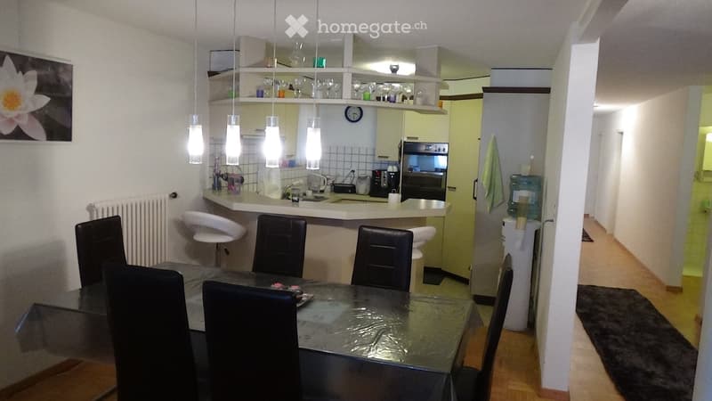 A modern, open-plan living and dining area with a sleek black dining table and chairs, a white kitchen counter with stools, and various appliances including an oven and refrigerator. The space features pendant lights, a radiator, and a water dispenser.