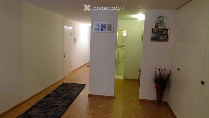 A well-lit hallway with wooden flooring, light-colored walls, and a small black rug on the left side. There are framed pictures on the walls and a vase with dried flowers on the right side. A doorway leads to a bathroom with white tiles and a towel visibl