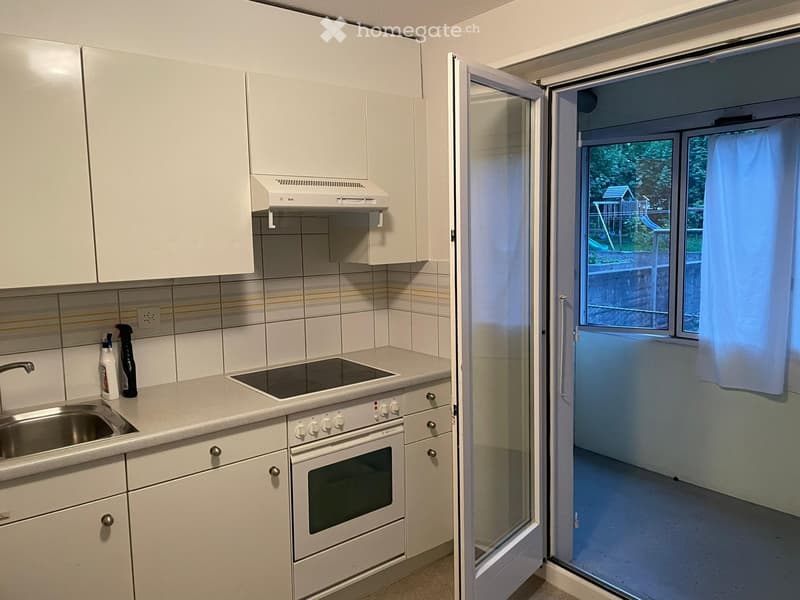 Modern kitchen with white cabinets, stainless steel sink, electric stove, and range hood. Open sliding door leads to a balcony with outdoor view.
