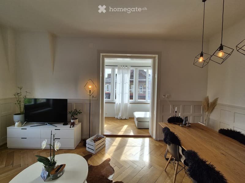 Bright living room with wooden flooring, large windows, modern furniture including a sofa, coffee table, chairs, and a TV on a white cabinet. A small bedroom is visible through an open doorway.