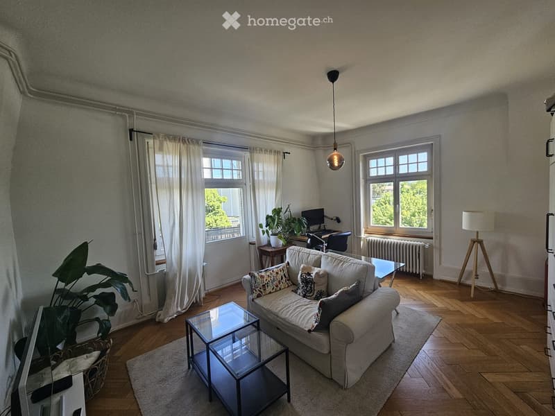 Living room with a light-colored sofa, coffee table, floor lamp, and a small desk with a computer. The room has wooden flooring, large windows with white curtains, and a potted plant near the window.