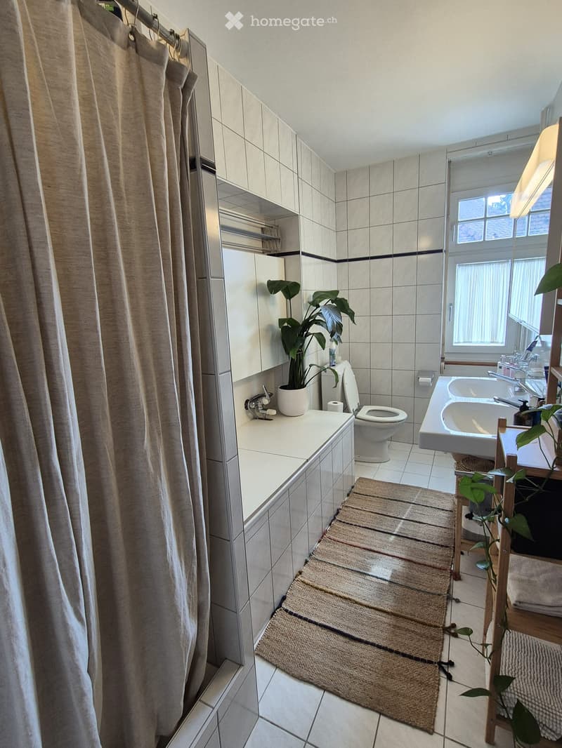 Modern bathroom with white tiles, a toilet, a sink, a bathtub, a shower curtain, and a potted plant on the counter.