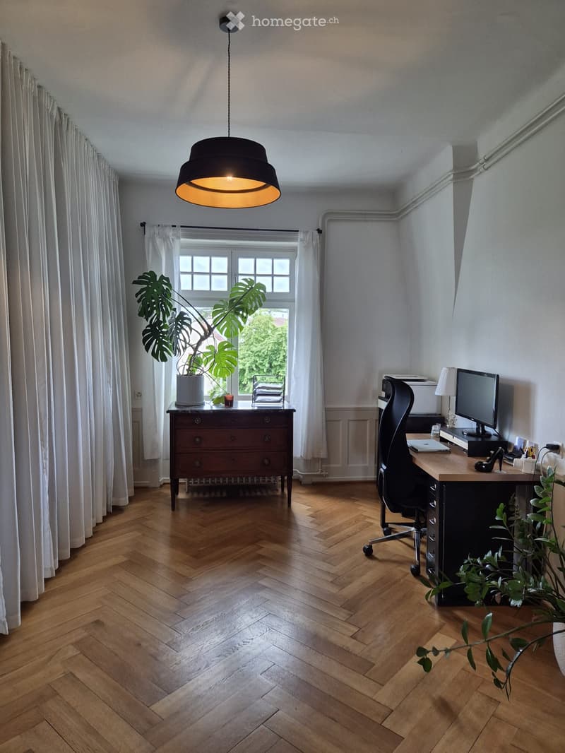 A well-lit, modern office space with wooden herringbone flooring, large windows, and contemporary furniture including a desk, chair, screen, and a hanging lamp.