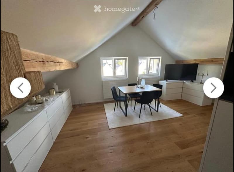 Attic room with wooden flooring, slanted ceilings, and exposed wooden beams. Features a dining area with a table and chairs, a television on a stand, and white cabinets along the walls.