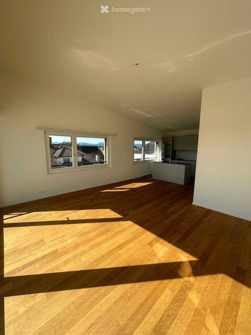 Empty room with light wooden flooring and large windows showing a view of rooftops and mountains. The room has white walls and a small kitchen area visible in the background.