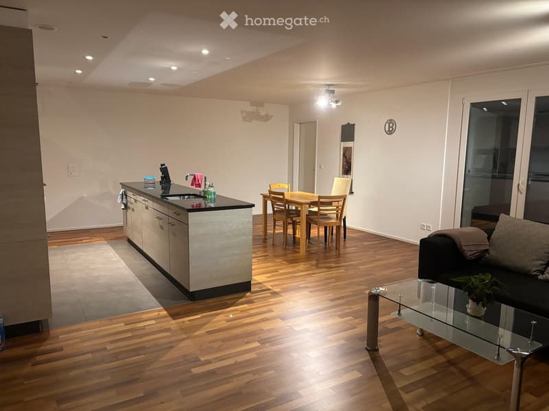 Open-plan living and dining area with wooden flooring, a black sofa, a glass coffee table, a dining table with chairs, and a kitchen island with a sink and screen.