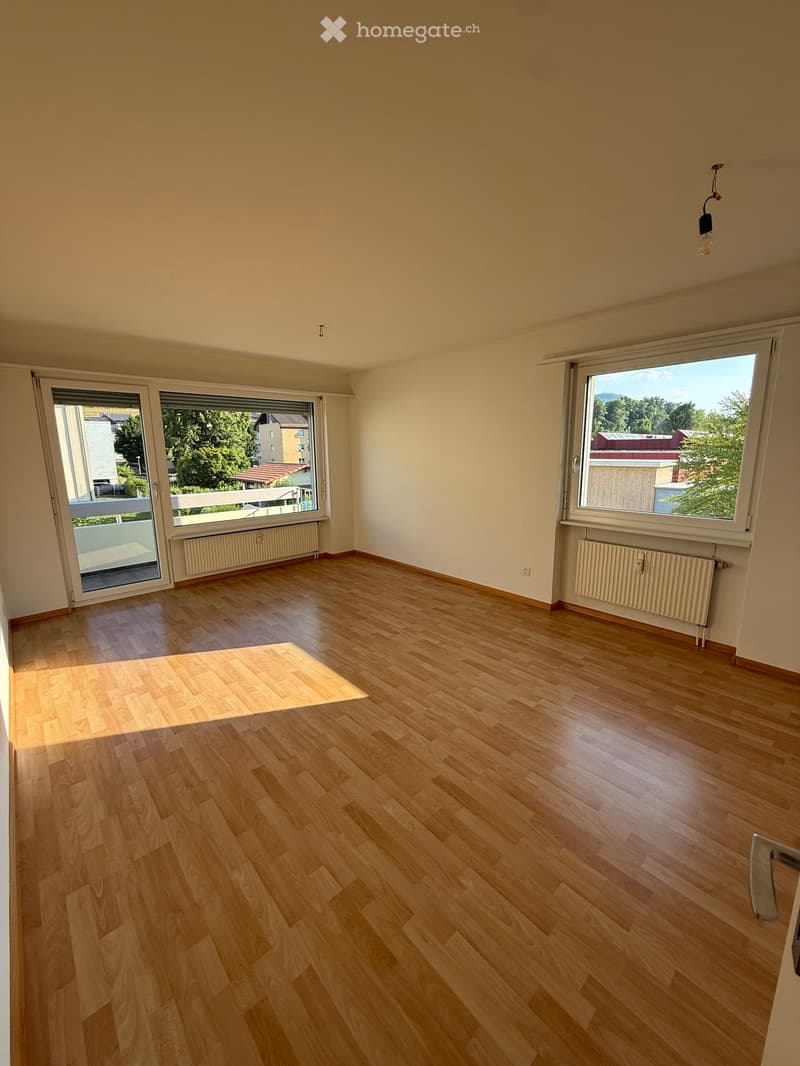 Empty room with wooden flooring, large windows, radiators, and a sliding glass door leading to a balcony.