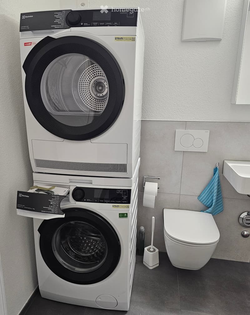 Modern bathroom with a stacked washing machine and dryer, toilet, sink, and a towel hanging on the wall.