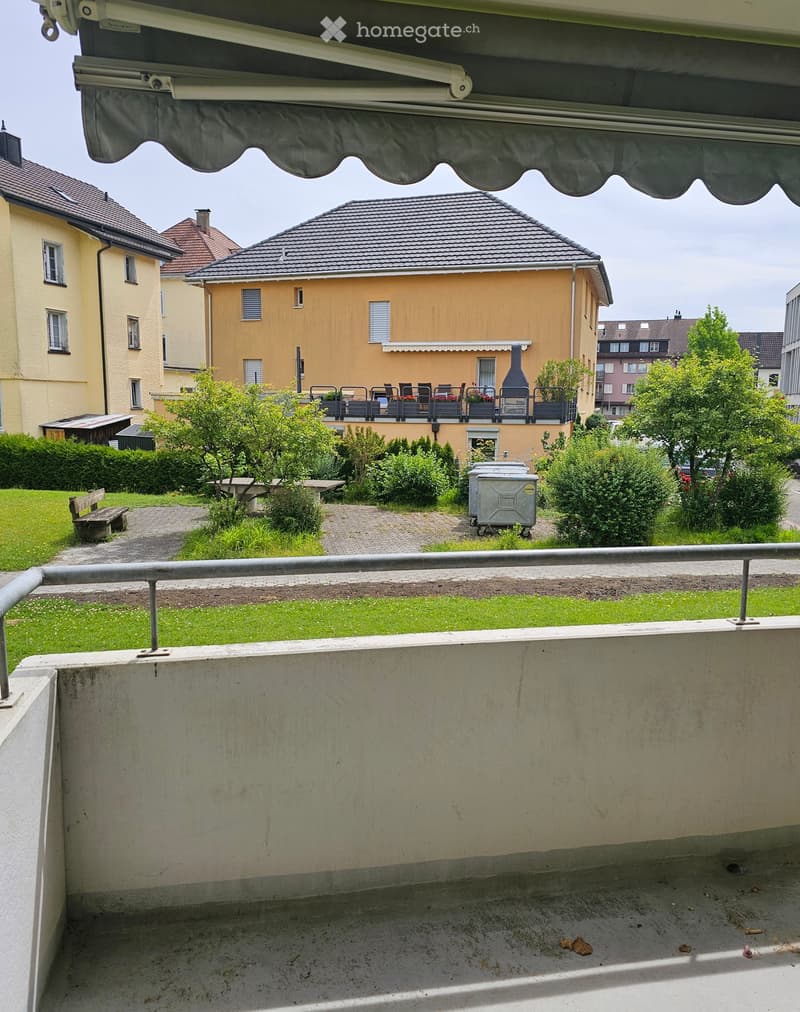 Empty balcony with concrete floor, metal railing, and view of a residential area with houses, greenery, and a covered patio area with outdoor furniture.