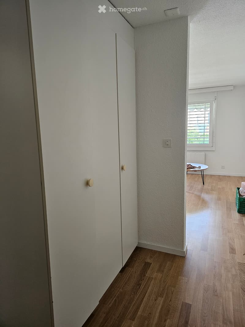 A small, well-lit room with wooden flooring, white walls, and a single window. A small table and chair are positioned near the window, and a green crate is visible on the floor.